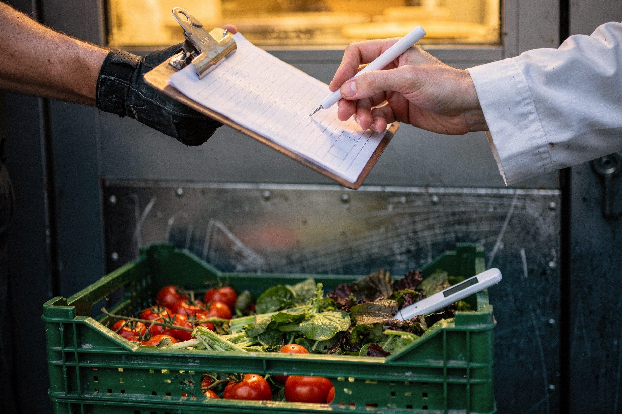 A delivery note being signed at a kitchen back door before dawn, with a probe thermometer resting in a crate of vine tomatoes and salad leaves